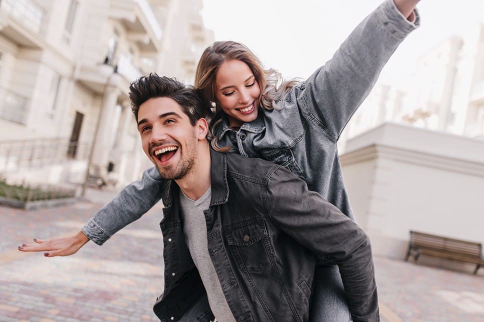 Un chico lleva a una chica en su espalda, ambos sonríen ella tiene los brazos abiertos