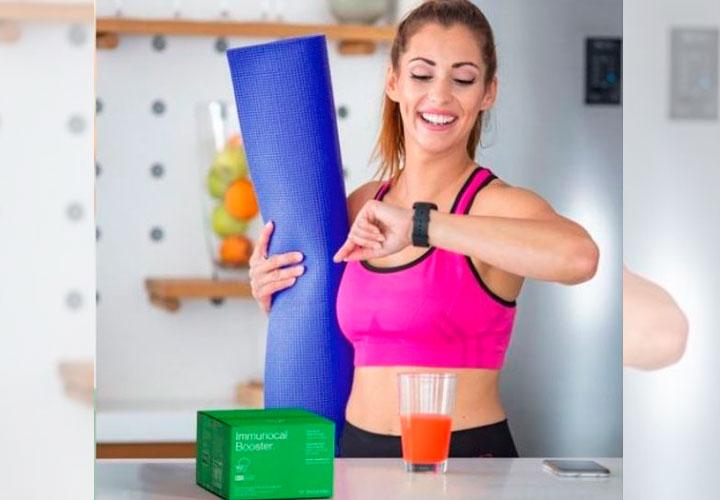 Chica mirando el reloj en una mano y en la otra lleva una esterilla. En la mesa un vaso de Immunocal booster verde preparado para tomar,